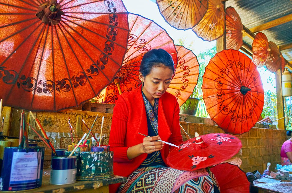 PINDAYA, MYANMAR - FEBRUARY 19, 2018:  The artisan of Burmese umbrella workshop creates the floral pattern for the small red parasol, on February 19 in Pindaya.
